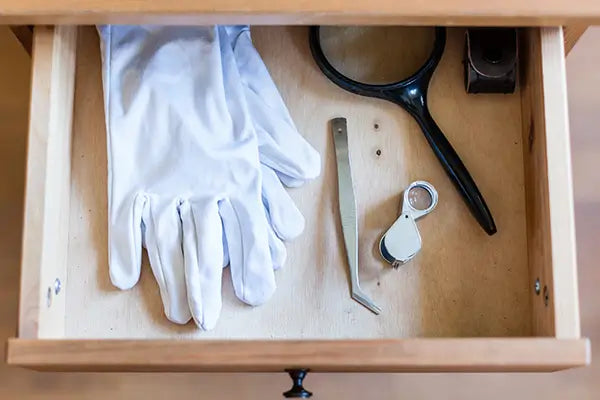 Une paire de gants blancs en coton est posée dans un tiroir ouvert avec une pince à épiler et une loupe.