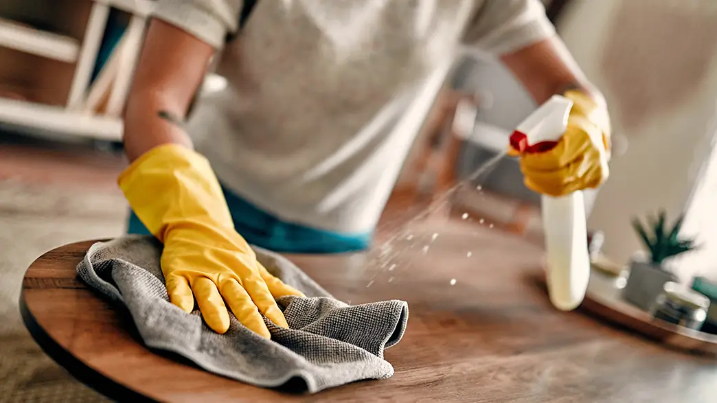 Eine Frau wischt die Oberfläche eines Holztisches feucht ab und benutzt dabei eine Sprühflasche mit Reinigungsmittel und trägt Schutzhandschuhe